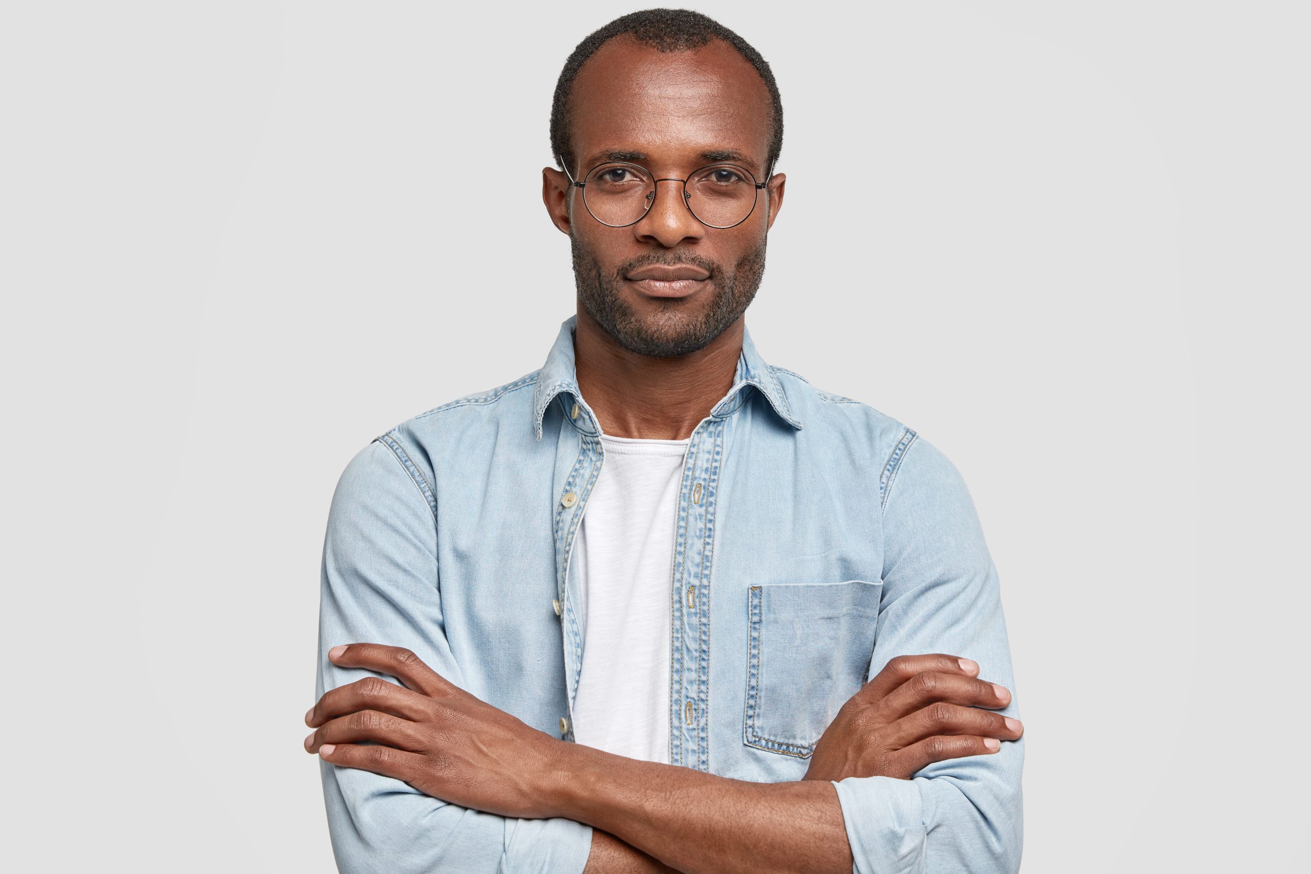 Serious confident male emterpreneur with dark healthy skin, keeps arms folded, listens attentively startup project, wears denim shirt, poses against white background. I am ready to listen you